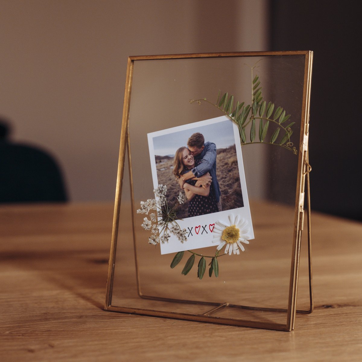 Polaroidka ve skleněném rámečku se zlatými detaily, ozdobená sušenými květinami.